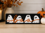 A rectangular black decorative sign with four white ghost shapes and an orange script word reading "boo" across the front.