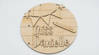 A round wooden sign with laser-cut details including stars, bunting, a pencil, and the text