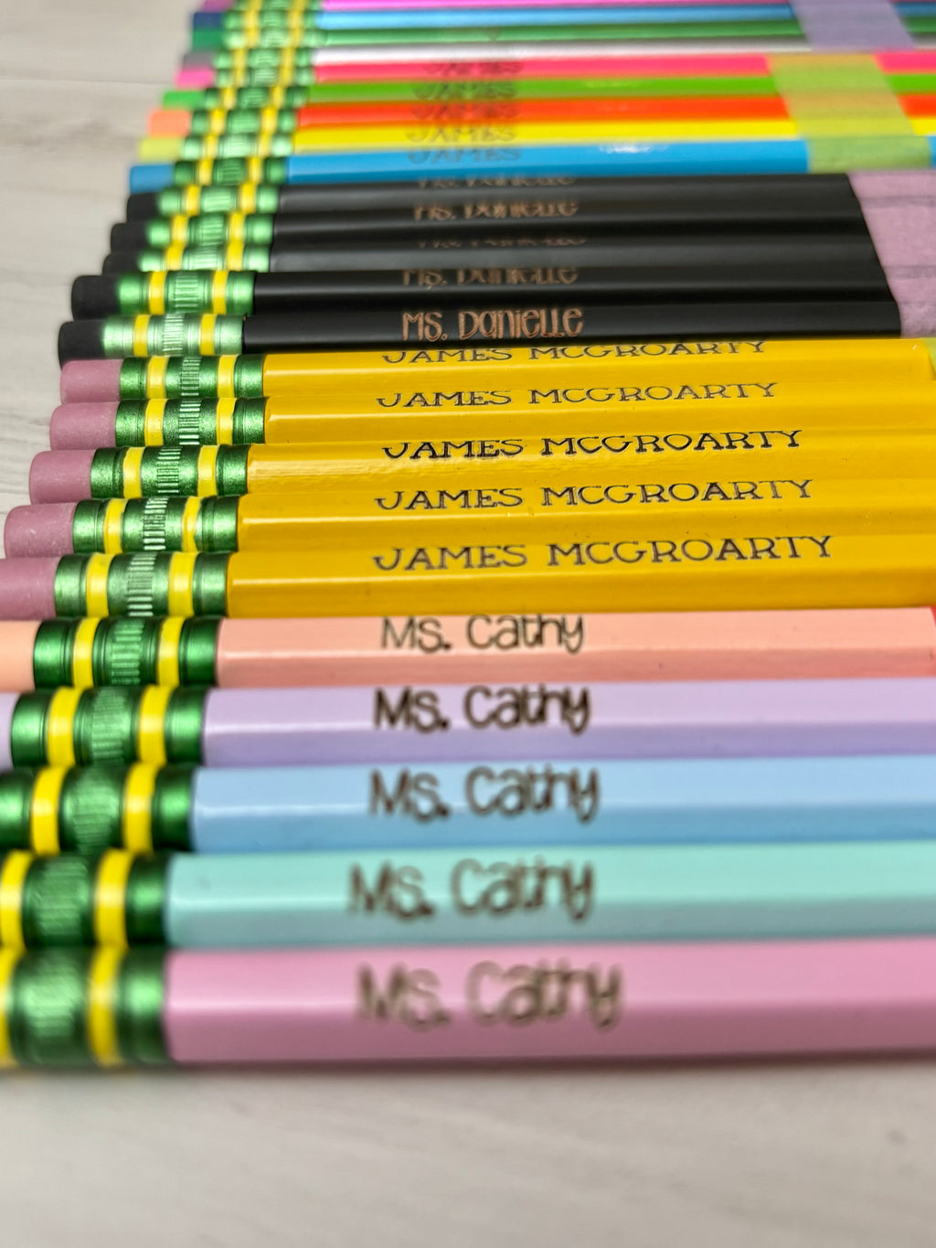 A close-up, angled view of multiple colored pencils arranged in rows. The pencils feature pastel pink, light blue, lavender, yellow, green, and black barrels, with engraved names on them. 