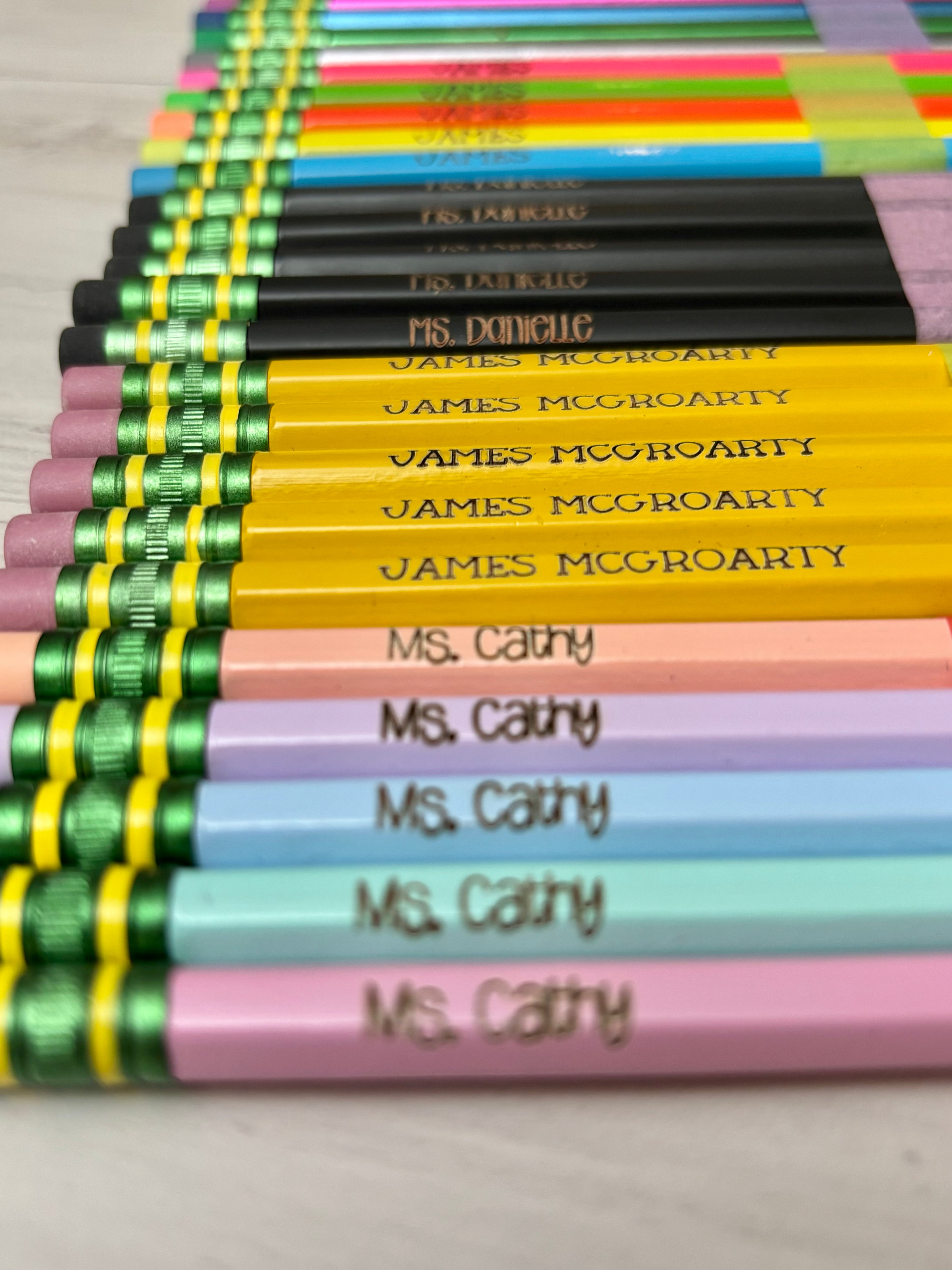 A close-up, angled view of multiple colored pencils arranged in rows. The pencils feature pastel pink, light blue, lavender, yellow, green, and black barrels, with engraved names on them. 