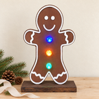 A gingerbread man shaped LED light on a wooden base, displayed on a tabletop with pine branches and pinecones. The light features a brown body with white outlines and glowing multicolor lights.
