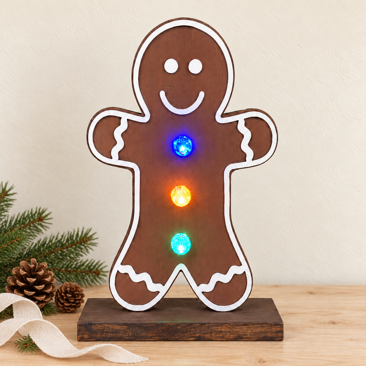 A gingerbread man shaped LED light on a wooden base, displayed on a tabletop with pine branches and pinecones. The light features a brown body with white outlines and glowing multicolor lights.