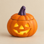 A carved jack-o'-lantern ceramic piece with a lid and a glowing warm light inside, featuring a purple stem, set against a solid beige background.