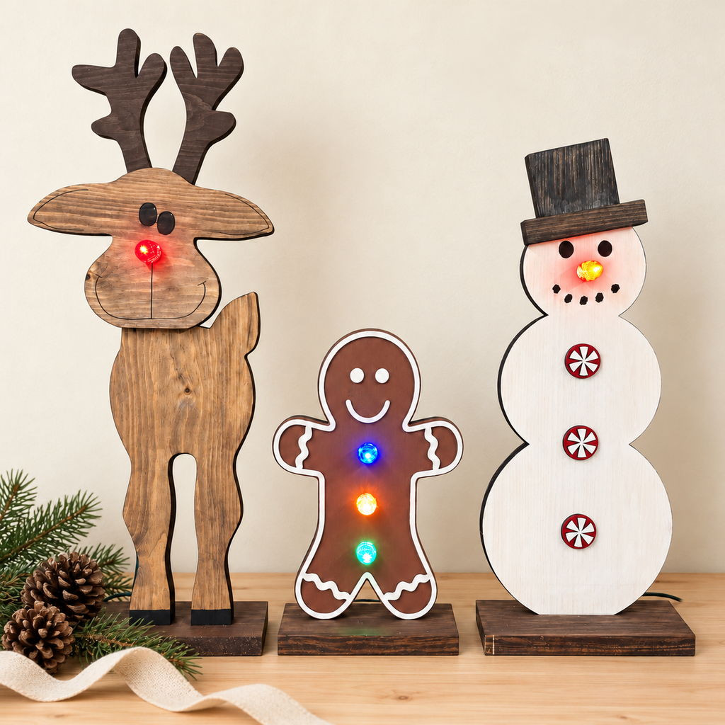Three wooden Christmas cutout figures on a tabletop: a reindeer with a red glowing light in its nose, a gingerbread man with multicolored glowing lights, and a snowman with red and yellow glowing lights. Pine branches and a ribbon are placed beside them against a plain beige wall.