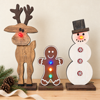 Three wooden Christmas cutout figures on a tabletop: a reindeer with a red glowing light in its nose, a gingerbread man with multicolored glowing lights, and a snowman with red and yellow glowing lights. Pine branches and a ribbon are placed beside them against a plain beige wall.