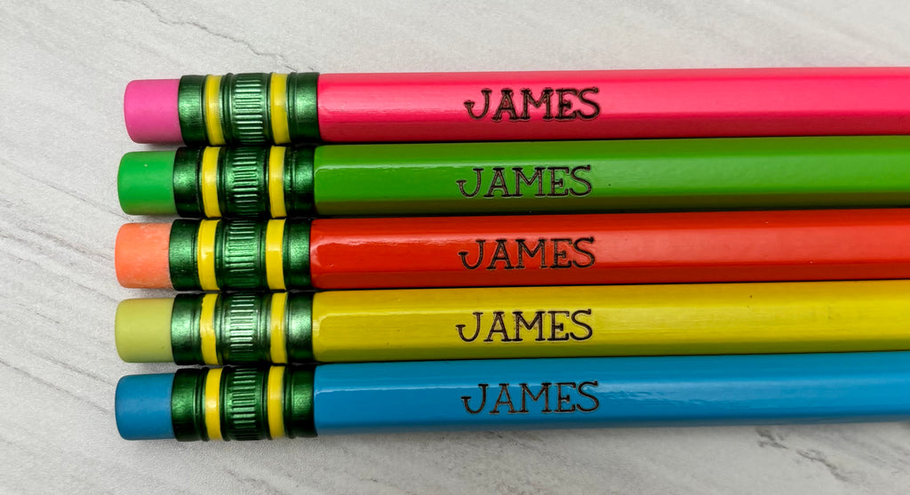 Five personalized wooden pencils in pink, green, orange, yellow, and blue, arranged horizontally. Each pencil is engraved.