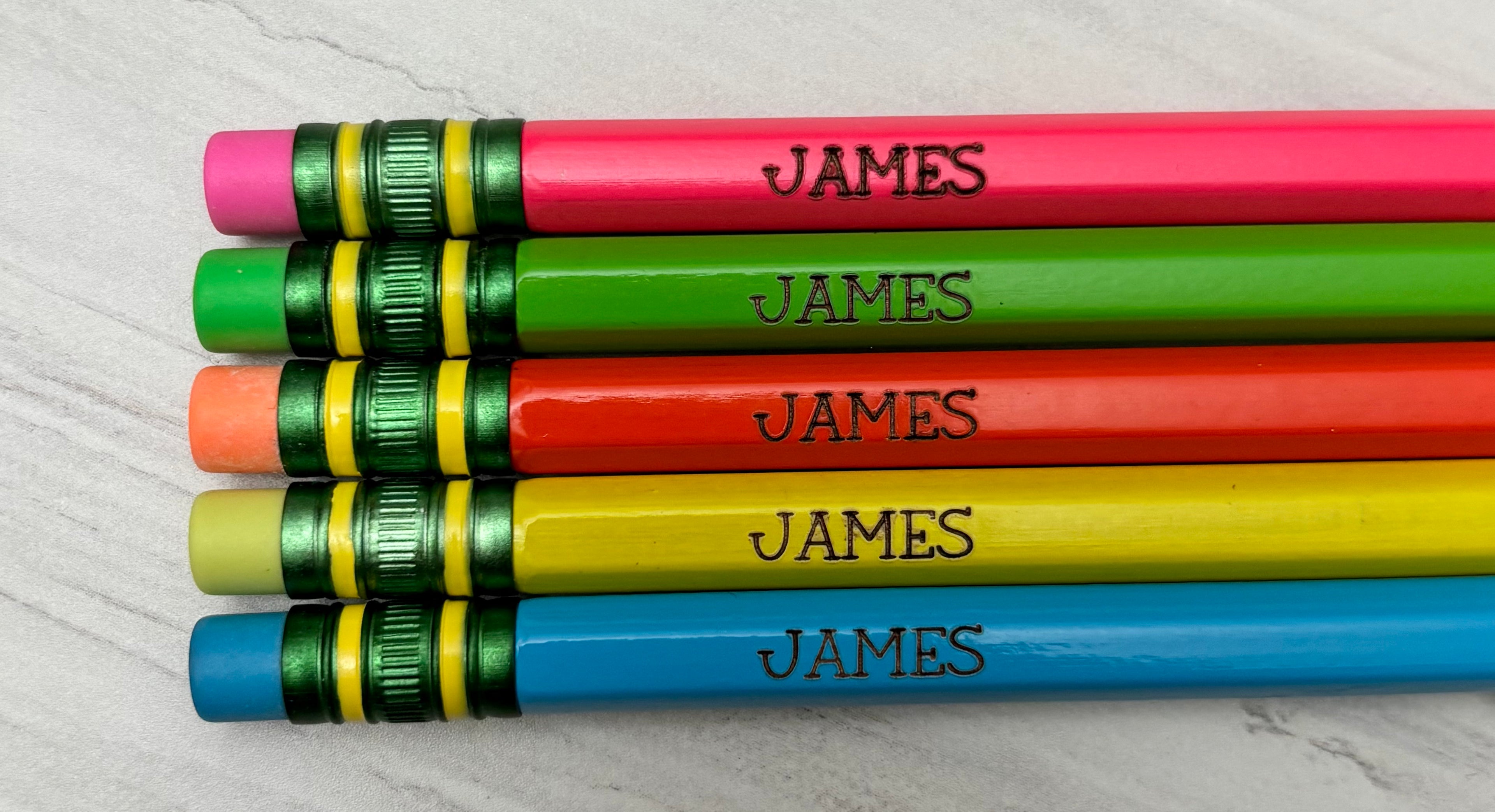 Five personalized wooden pencils in pink, green, orange, yellow, and blue, arranged horizontally. Each pencil is engraved.
