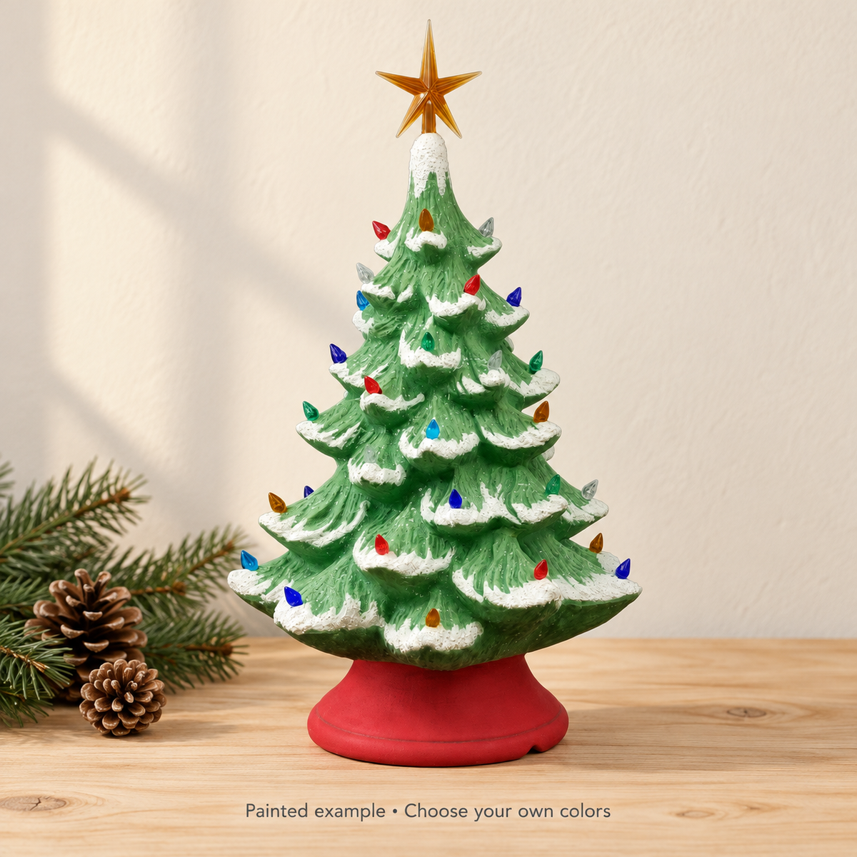 A ceramic Christmas tree with a red base, green branches, white snow-like tips, multicolored lights, and a gold star on top, displayed on a wooden surface with pine branches and pinecones beside it. Text at the bottom reads 'Painted example • Choose your own colors'.