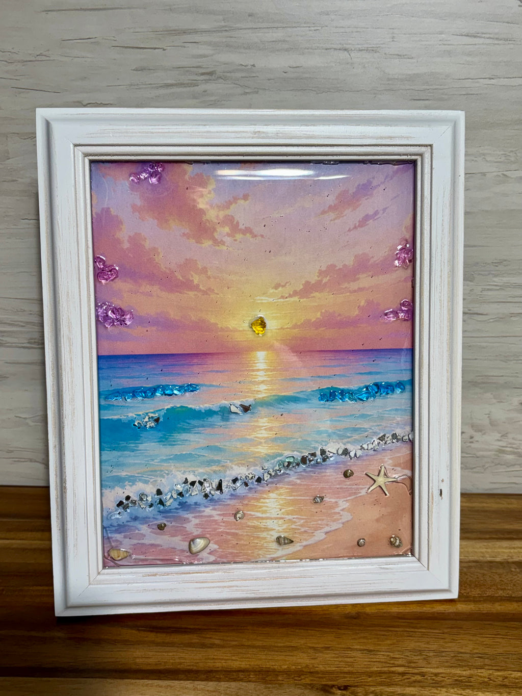 Framed painting of a sunset over the ocean accentuated with glass and shells.