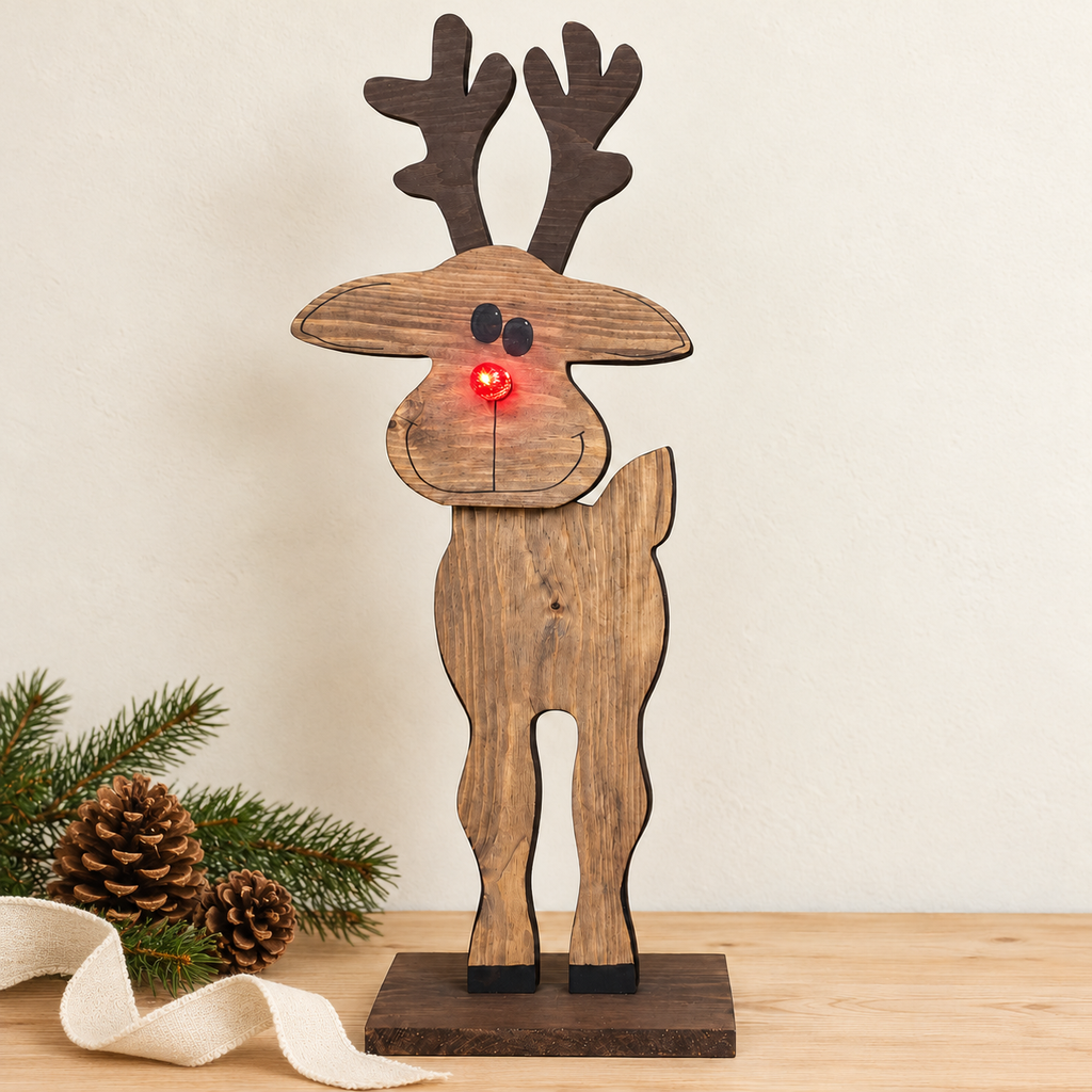 A rustic wooden Christmas reindeer figurine with dark brown antlers and face, medium brown wood-grain body, black eyes and hooves, and a glowing red nose, standing on a dark base on a tabletop with pine branches and a ribbon nearby.