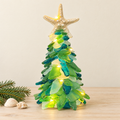 A decorative Christmas tree made from layered translucent green and blue sea glass pieces, lit with warm white fairy lights, topped with a glittery gold starfish. The tree is styled on a light wood surface with small seashells and a pine branch against a neutral beige background.