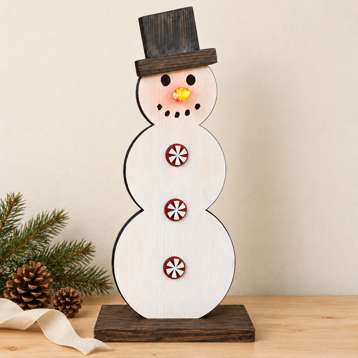 A wooden snowman tabletop decoration with three stacked oval sections, a dark top hat, red-and-white candy cane buttons, and a small yellow nose, displayed on a wooden surface with pine branches and pinecones at the side.