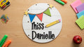 A personalized round white sign with black script, decorated with colorful bunting, stars, and a yellow pencil.
