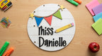 A personalized round white sign with black script, decorated with colorful bunting, stars, and a yellow pencil.