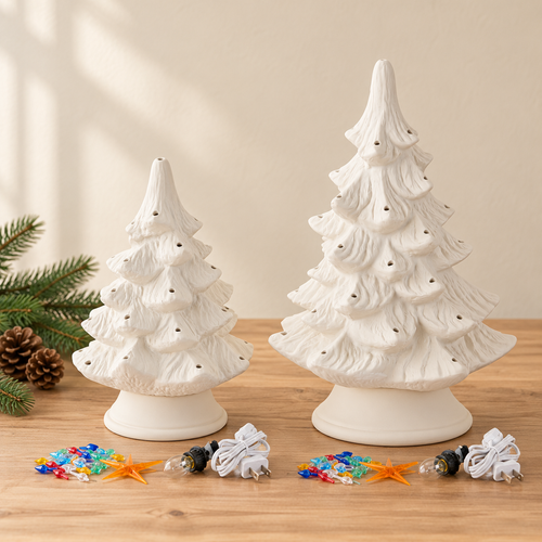 Two unpainted ceramic Christmas trees on a wooden table, with small colorful lights and a star topper placed in front. A pine branch and pinecone are visible at the left edge against a warm beige wall.